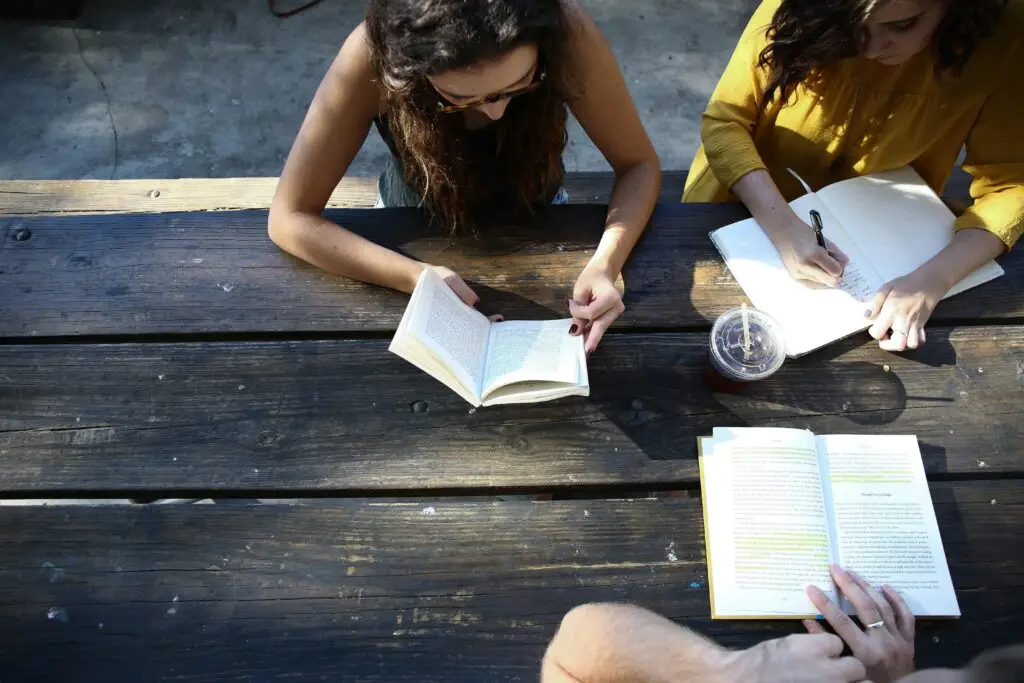 Three people studying together outdoors at wooden table with books and compass.