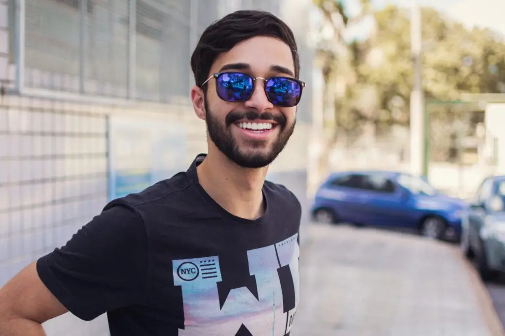Smiling man wearing sunglasses and NYC graphic t-shirt outdoors on sunny day.