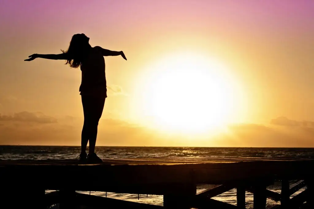 Silhouette of woman with arms outstretched on pier at sunset over ocean.