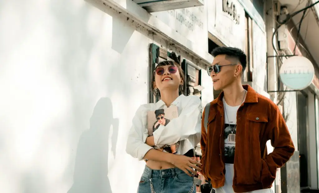 Two young people wearing sunglasses stand together against a white building wall.