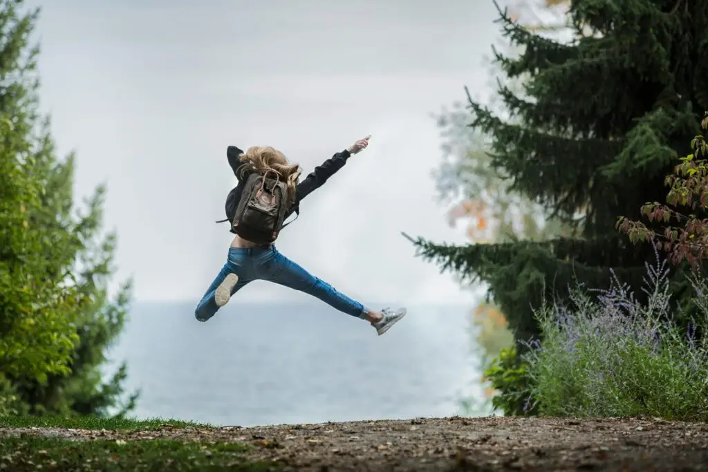 Woman jumping joyfully outdoors surrounded by evergreen trees and water.