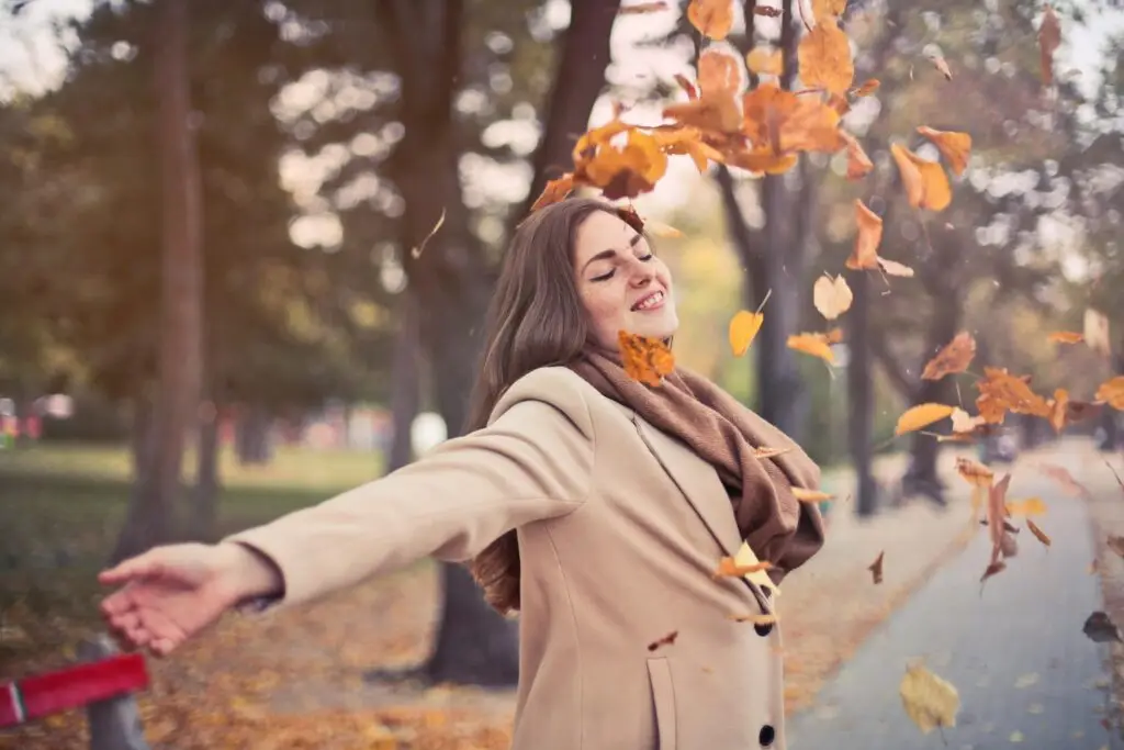 Woman in beige coat enjoying autumn leaves falling in park during fall season.