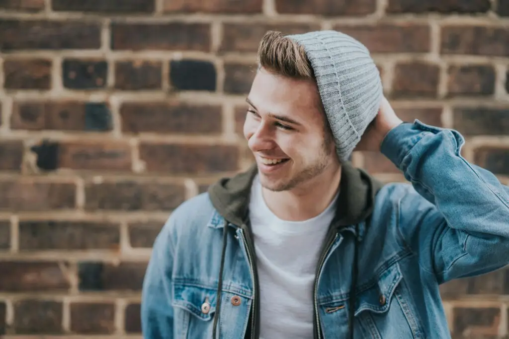 Young man in denim jacket and beanie smiling against brick wall.