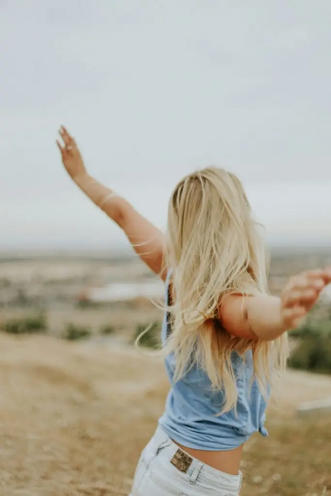 Woman with blonde hair stands on overlook with arms raised toward sky in celebration.
