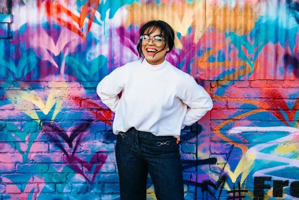 Smiling young person in white sweater standing against vibrant colorful graffiti wall.