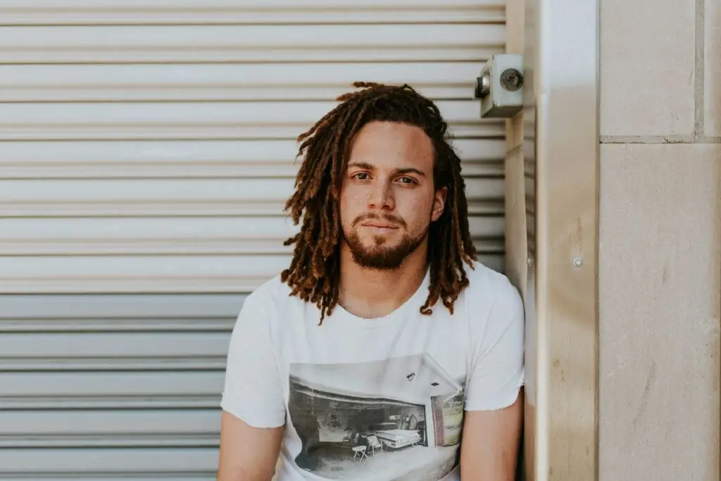 Man with dreadlocks wearing white graphic t-shirt against corrugated metal wall.