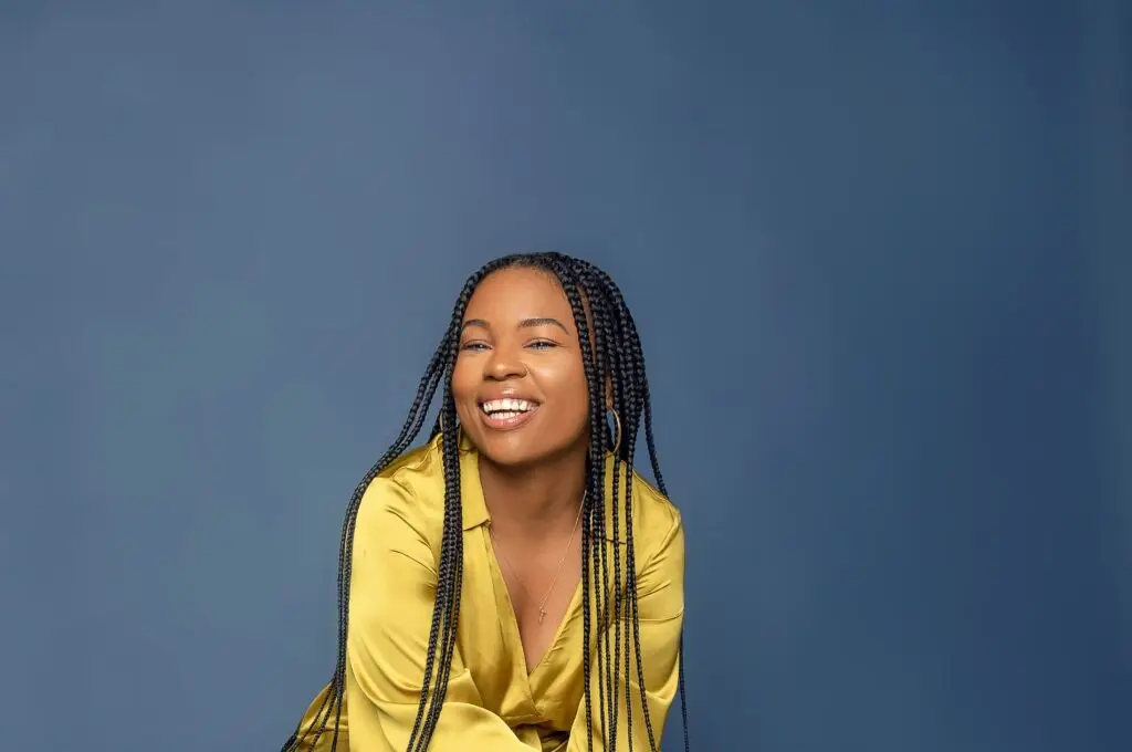 Smiling woman with braided hair wearing yellow shirt against blue background.