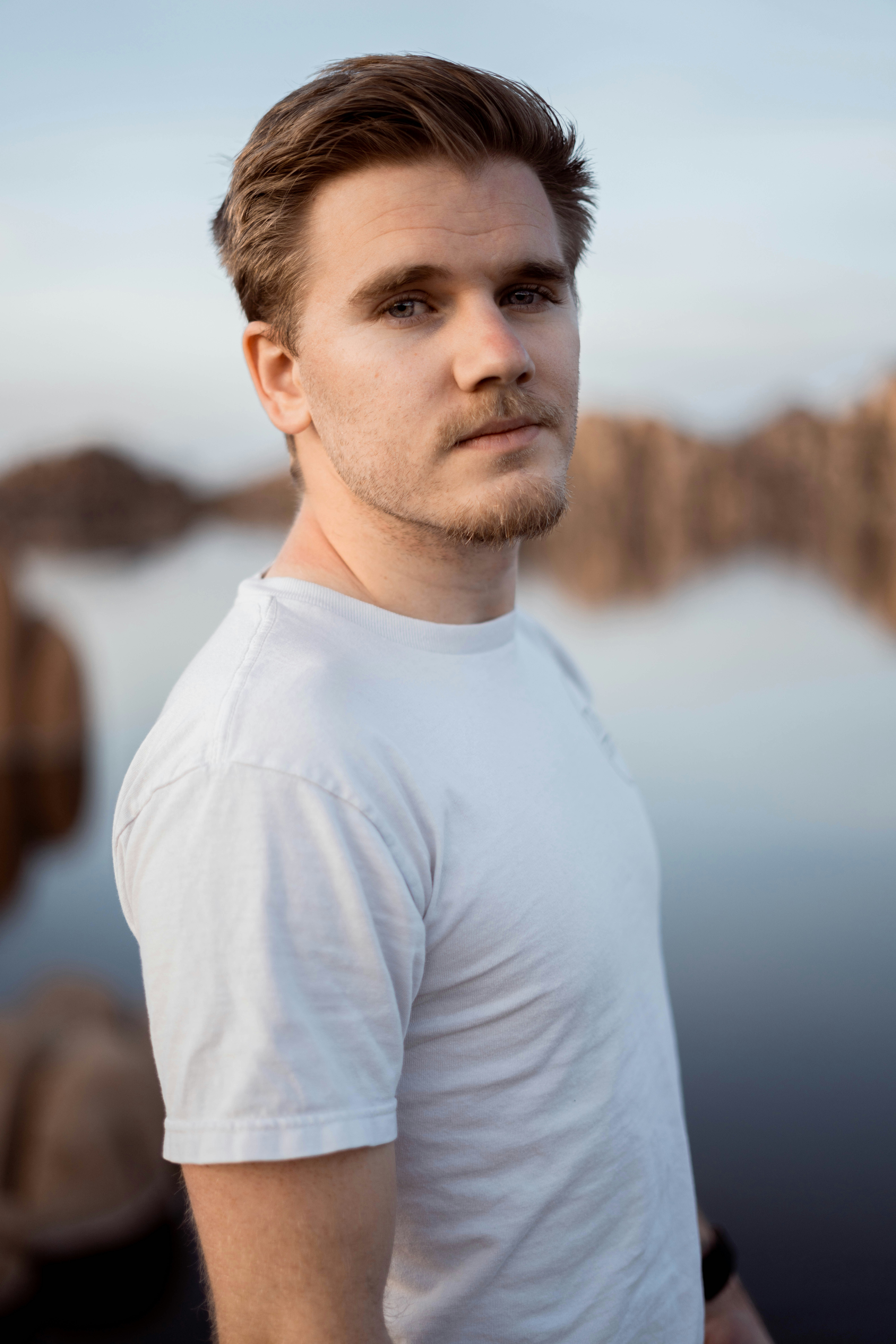 Man in light blue shirt standing outdoors by water, looking thoughtful.