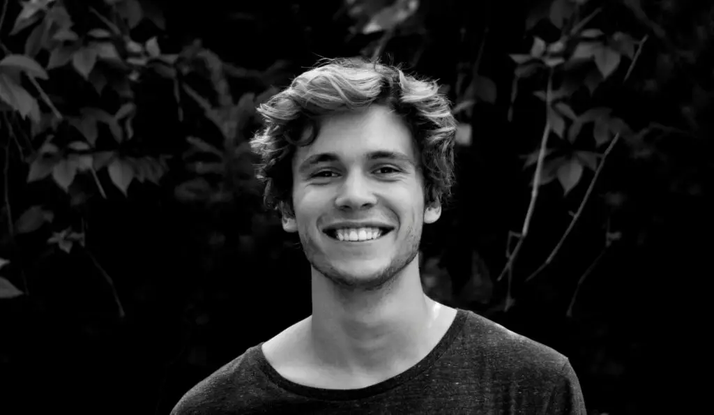Smiling man in casual shirt against dark foliage background, portrait style photograph.