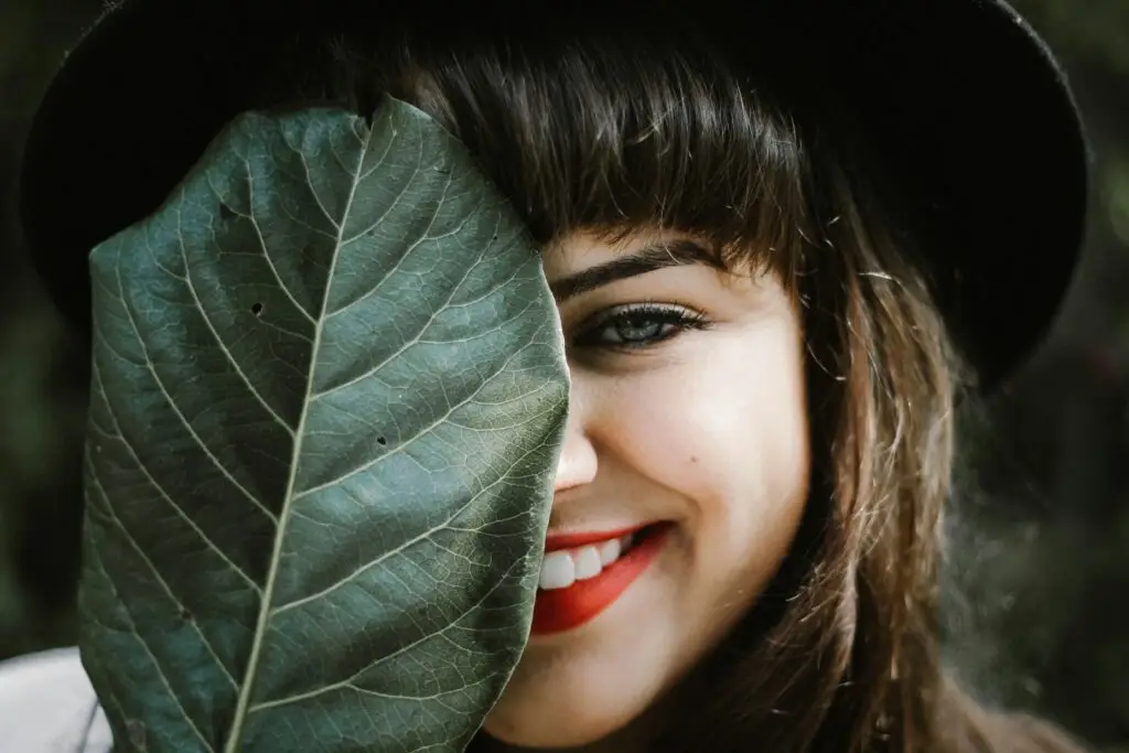 Woman holding large green leaf, smiling with red lipstick and dark hair.