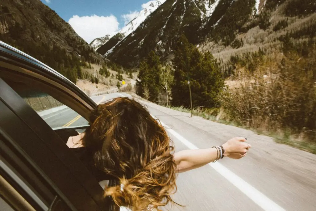 Woman with long brown hair leaning out car window toward mountain valley scenery.