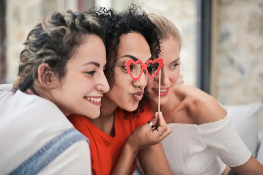 Three smiling women holding heart-shaped glasses together indoors.
