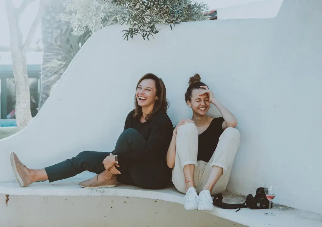 Two young women sitting against white wall, laughing together outdoors.