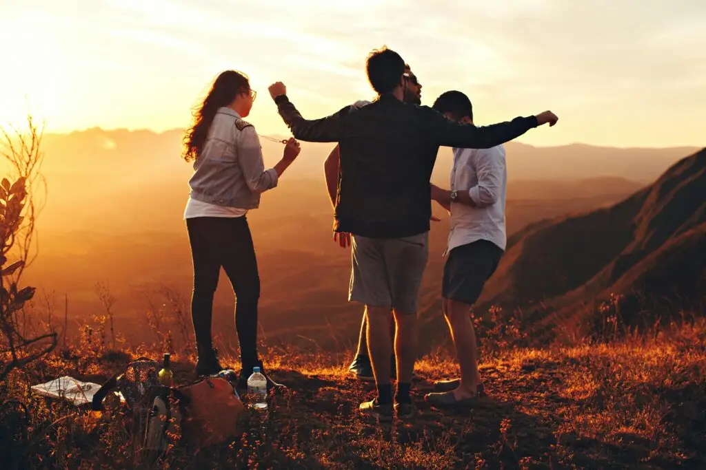 Three people celebrating on a mountain overlook at sunset with arms raised.