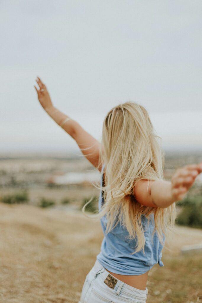 Woman with blonde hair standing in open field with arms raised in freedom pose.