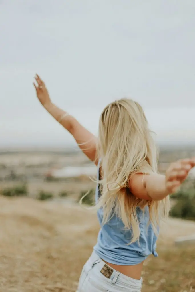Woman with blonde hair standing in open field with arms raised in freedom pose.