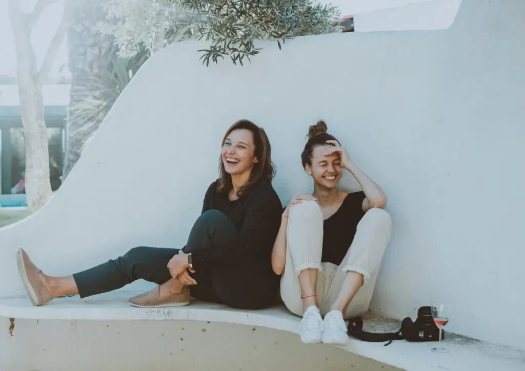 Two women sitting together against white wall, laughing and smiling outdoors.