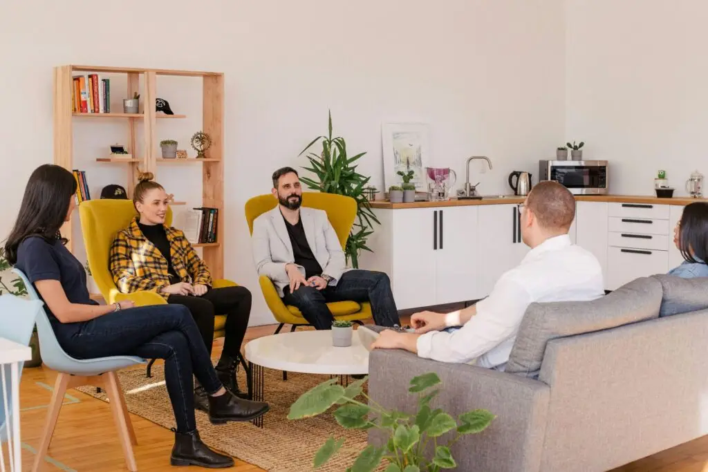 Five adults in group therapy session sitting in modern, plant-filled office space.